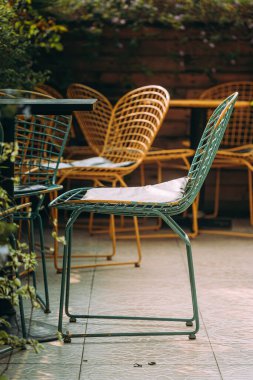 Kablodan yapılmış hasır sandalyeler fayansta bir ağacın yanında duruyor. Orada kimse yok, sıradışı sandalyeler bahçede duruyor. Metal bacaklı koltuklar. Nostaljik ve barışçıl hissediyorum. Seçici odak.