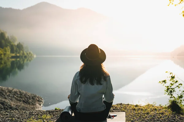 年轻的女徒步旅行者 戴着帽子 穿着牛仔夹克 独自在日出山中冥想瑜伽 从后面看旅行生活方式精神放松的概念 与自然和谐相处 — 图库照片