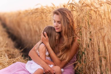 Anne, altın buğday tarlasının yanında dizlerinin üzerinde oturan küçük kızını nazikçe kucaklıyor. Genç bir kadın bir kızı sevgiyle okşar. Kızlar çim biçme tarlasının yanında oturuyorlar. Hafta sonu kır yürüyüşleri.