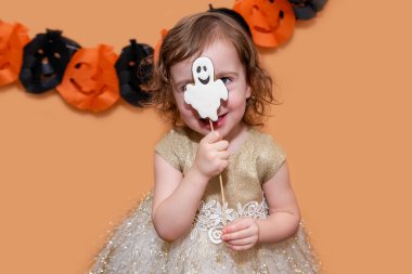 Küçük kız hayalet şeklinde zencefilli kurabiyelerle eğleniyor, izole bir arka planda siyah ve turuncu balkabağı desenli bir çelenkle suratını buruşturuyor. Cadılar Bayramı hediyesi. Boşluğu kopyala