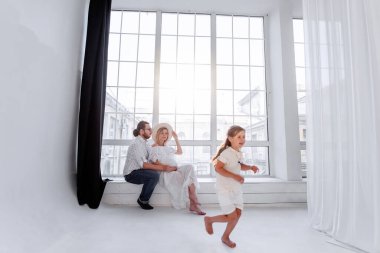 Mutlu aile, panoramik pencerenin yanındaki pencere eşiğinde otururken eğleniyor. Genç kadın hamile, erkek sarılıyor. Küçük kız anne babasıyla oynaşıyor, dans ediyor. Minimalist tavan arası.