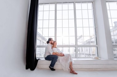 Genç adam hamile kadının karnına sarılıyor. Sevgili çift, bebeğin doğumu beklentisiyle panoramik pencerenin kenarındaki tahta pencere eşiğine oturur. Mutlu, sağlıklı bir aile. Hamilelik sırasındaki koca desteği