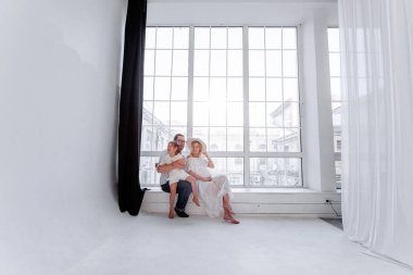 Mutlu aile, panoramik pencerenin yanındaki pencere eşiğinde otururken eğleniyor. Genç kadın hamile, erkek sarılıyor. Küçük kız anne babasıyla oynaşıyor, dans ediyor. Minimalist tavan arası.