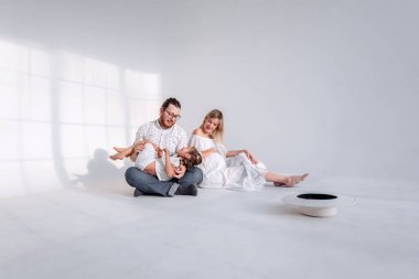 Beyaz minimalist iç mekanda, aile yerde oturarak eğleniyor. Genç sarışın hamile kadın gülüyor, kızına ve kocasına bakıyor. Kız babasıyla takılıyor, koşuyor.