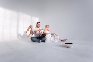 Beyaz minimalist iç mekanda, aile yerde oturarak eğleniyor. Genç sarışın hamile kadın gülüyor, kızına ve kocasına bakıyor. Kız babasıyla takılıyor, koşuyor.