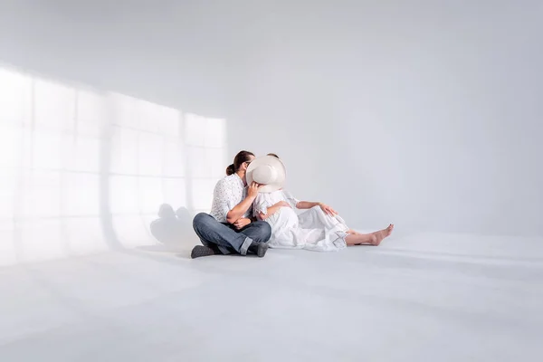 若い女性は妊娠しており 男性は帽子で顔を覆い 女の子にキスをします 白いミニマリストスタジオのカップルを愛する床に座っている 幸せな家族 健康な妊娠 背景の光影 — ストック写真