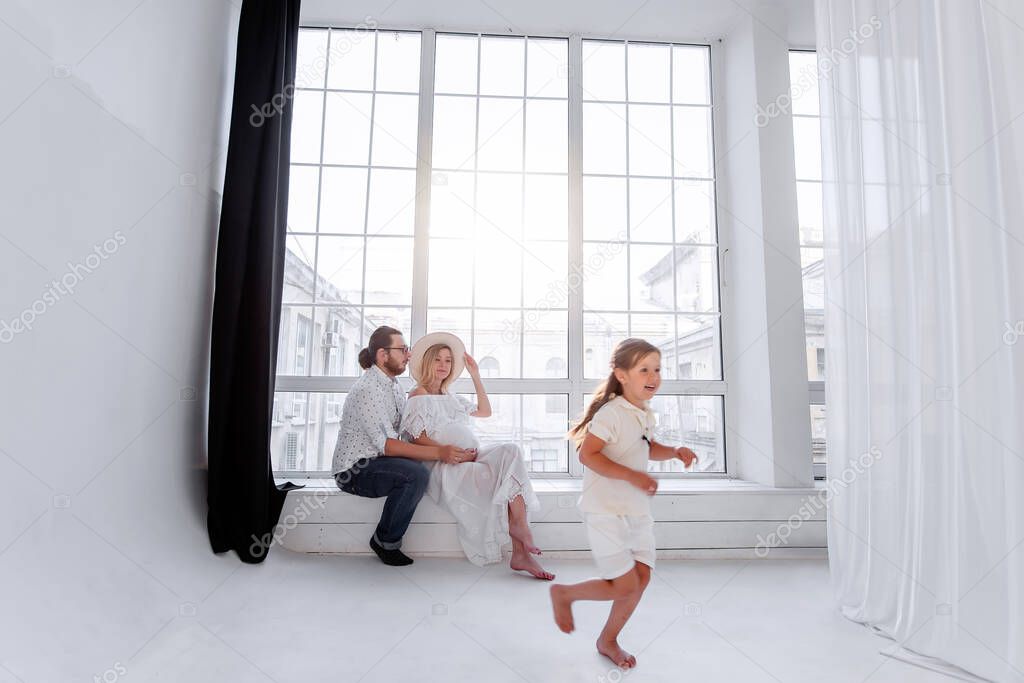 Happy family is having fun while sitting on the windowsill by the panoramic window. Young woman is pregnant, man hugs beloved. Little girl fooling around parents, dancing. Minimalist loft interior