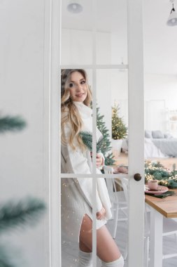 Modern mutfakta, Noel ağaçlarının arasında şık iç mekanlarda, uzun beyaz, örgü süveter giymiş genç bir kadının moda portresi. Dolabın camından bakan kıvırcık sarışın kız.