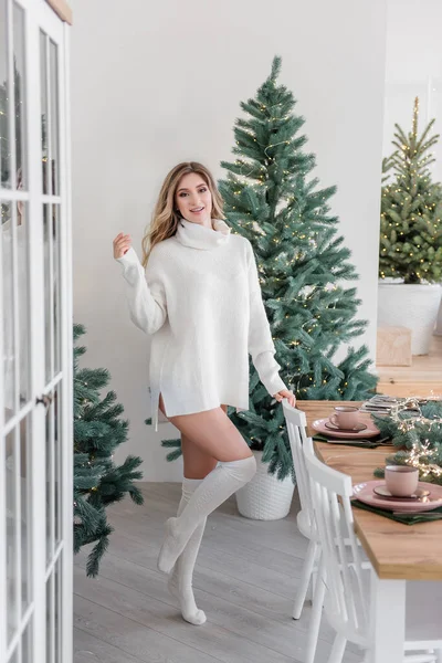 長い白いニットセーターのドレスを着たファッションの若い女性は レギンスは ファッショナブルなインテリアのクリスマスツリーの中で現代的なキッチンで踊って 楽しんでいます お祝いの新年の雰囲気の中で現在ブロンドの女の子 — ストック写真