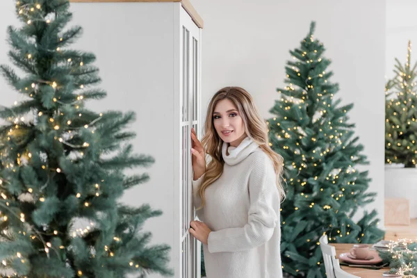 長い白いニットセーターのドレスを着たファッションの若い女性は レギンスは ファッショナブルなインテリアのクリスマスツリーの中で現代的なキッチンで踊って 楽しんでいます お祝いの新年の雰囲気の中で現在ブロンドの女の子 — ストック写真
