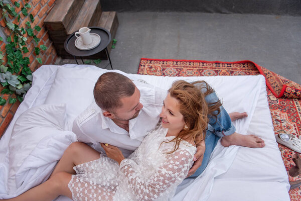 Loving couple of millennials hug each other, resting on white bed against background of brick wall with green ivy. Man passionately embraces young woman, kisses gently. Lovers weekend in the morning