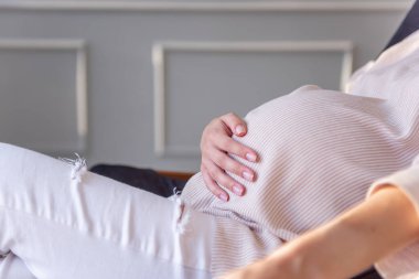 Genç hamile bir kadının portresi ahşap bir iskemlede oturuyor ve karnına sarılıyor. Mutlu annelik, sağlıklı hamilelik. Bir bebeğin doğacağı beklentisiyle, elinde bir şapka tutan moda kızı.