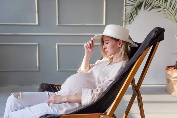 木製のデッキチェアに座っているスタイリッシュな妊婦の肖像画 頭の上に白い帽子をかぶって 幸せな母親 健康な妊娠 赤ちゃんの誕生を期待してファッションの女の子 トレンディーなロフトインテリア — ストック写真