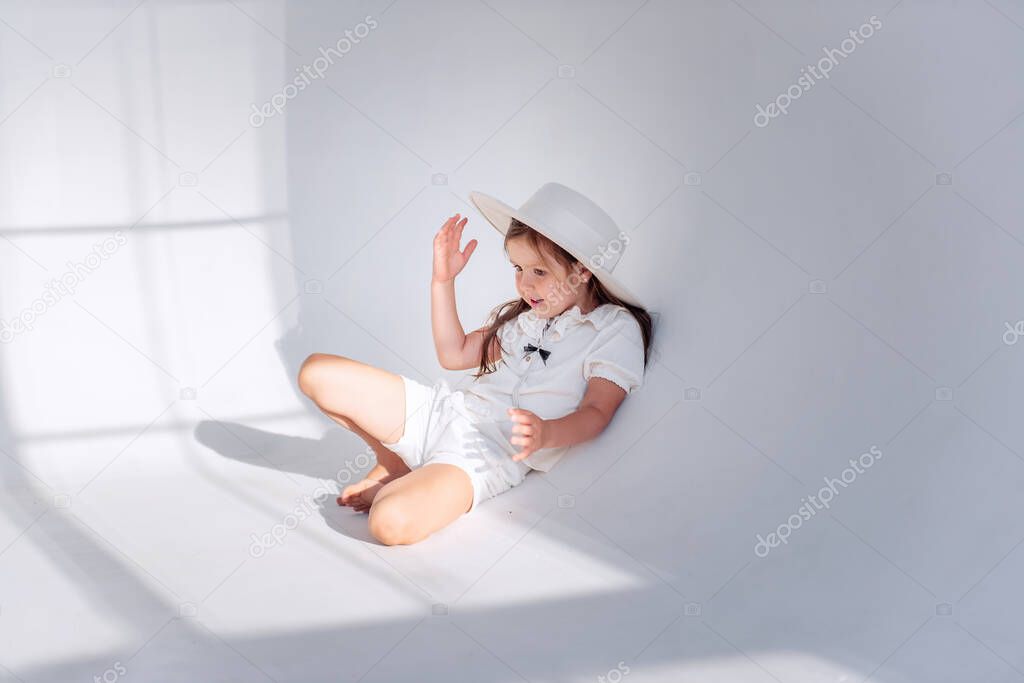 Little girl is having fun on the floor in stylish hat in a white loft interior with airy fabrics. Shadows from the window on the floor, silhouette of a child. Cheerful childhood, the girl grimaces