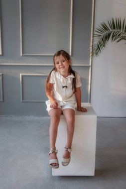 Küçük bir kız beyaz takım elbiseli, saçları açık bir küp üzerinde oturuyor. Moda çocuğu, yeşil palmiye ağacıyla gri arka planda model bir okulda poz veriyor.