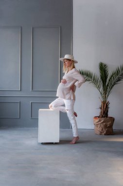 Beyaz şapkalı hamile bir kadın gri, şık bir stüdyoda bacağını beyaz bir küpe yaslıyor. Kotlu kız karnına şefkatle sarılıyor. Güzel, sağlıklı hamilelik.