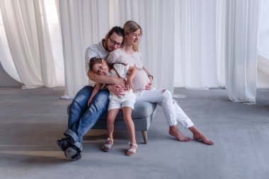 Minimalist stüdyoda, hamile genç bir kadın bir puf üzerinde oturur, sakallı bir adam küçük bir kıza sarılır. Çocuk beklentisi içinde mutlu bir aile. Baba anneye sarılır, kız güler. Sağlıklı hamilelik