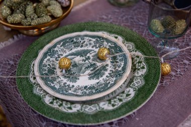 Mor bir masa örtüsünün üzerinde eski yeşil ve beyaz tabaklar ve süsler vardı. Altüst olmuş altın toplardan oluşan bir çelenk vardı. Kutlama masası. Kozalaklı Noel süslemesi. Boşluğu kopyala