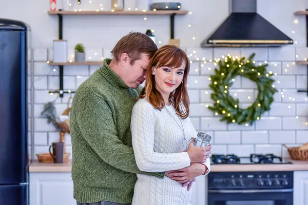 クローズアップの肖像ニットのセーターを着た素敵なカップルは スタイリッシュなインテリアのファッショナブルなキッチンで抱き合っています 北欧スタイル クリスマスの装飾 スラブの上に緑のトウヒの花輪がかかっています — ストック写真