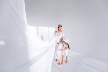 Hamile anne minimalist iç kısımda beyaz hafif kumaş tutuyor. Pencereden yansıyan hafif gölgeler var. Şapkalı küçük kız etrafta koşuşturuyor, eğleniyor. Sağlıklı hamilelik, mutlu aile.