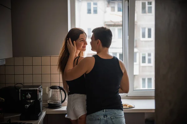 年轻的两千年夫妇穿着黑色的T恤衫 早上在厨房里抱着牛仔裤 挨着窗户 爱你的人在回家吃早饭前会玩得很开心 男人抚摸女人的头发 生活方式的公寓生活 — 图库照片