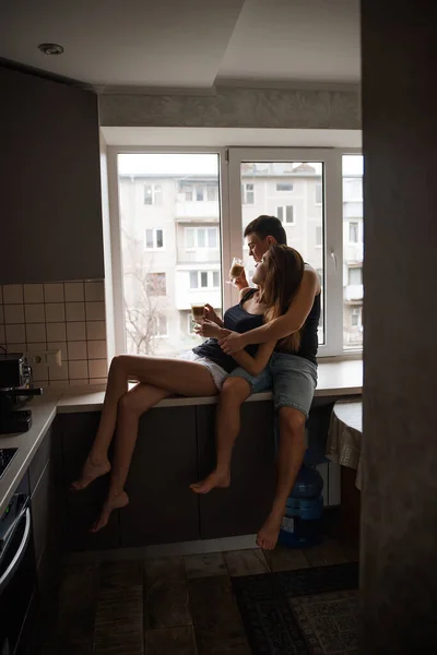 若い千年のカップルが窓の近くの窓の上に座っていて アパートの自宅のキッチンでラテコーヒーを飲んでいます 恋人抱擁 楽しみを持っています ガラスコップを持っている男と女 — ストック写真