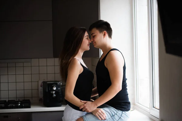 年轻的两千年夫妇穿着黑色的T恤衫 早上在厨房的窗边抱着牛仔裤 爱你的人在回家吃早饭前会玩得很开心 男人坐在窗台上 亲吻站着的女人 — 图库照片