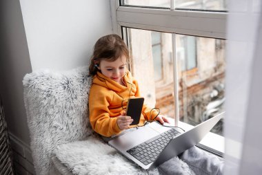 Küçük kız geniş pencere pervazında oturur, dizüstü bilgisayarda oyunlar oynar, kulaklıkla müzik dinler, akıllı telefondan selfie çeker. Pencerenin yanındaki turuncu kapüşonlu gri pantolonlu çocuk. Çocuklar Teknolojiyi Kullanıyor