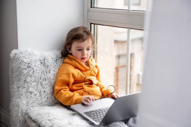 Küçük kız geniş pencere pervazında oturur, dizüstü bilgisayarda oyunlar oynar, kulaklıkla müzik dinler. Turuncu kapüşonlu çocuk, evin penceresinde gri pantolon. Anaokulu öğrencileri için uzaklık öğrenme