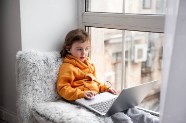 Küçük kız geniş pencere pervazında oturur, dizüstü bilgisayarda oyunlar oynar, kulaklıkla müzik dinler. Turuncu kapüşonlu çocuk, evin penceresinde gri pantolon. Anaokulu öğrencileri için uzaklık öğrenme