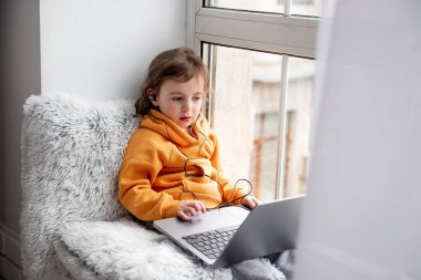 Küçük kız geniş pencere pervazında oturur, dizüstü bilgisayarda oyunlar oynar, kulaklıkla müzik dinler. Turuncu kapüşonlu çocuk, evin penceresinde gri pantolon. Anaokulu öğrencileri için uzaklık öğrenme