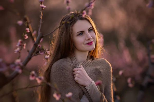 Young millennial woman in pink blooming sakura gardens at sunset. Close-up portrait of natural beauty girl in tender peach blossoms. Walks outside the city, the countryside at the weekend. Copy space