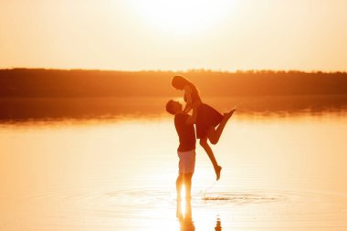 Silhouette of couple in love, dances with splashes in the water of the lake in the rays of orange sunset. Lovers hug each other, kiss. The man circles the woman in arms. Honeymoon travel of newlyweds