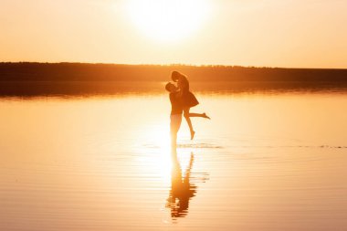 Silhouette of couple in love, dances with splashes in the water of the lake in the rays of orange sunset. Lovers hug each other, kiss. The man circles the woman in arms. Honeymoon travel of newlyweds