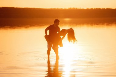 Silhouette of couple in love, dances with splashes in the water of the lake in the rays of orange sunset. Lovers hug each other, kiss. The man circles the woman in arms. Honeymoon travel of newlyweds