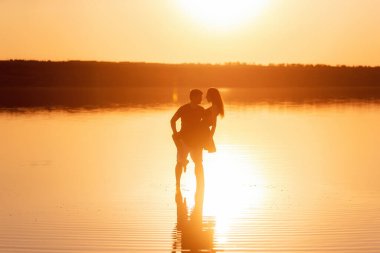Silhouette of couple in love, dances with splashes in the water of the lake in the rays of orange sunset. Lovers hug each other, kiss. The man circles the woman in arms. Honeymoon travel of newlyweds