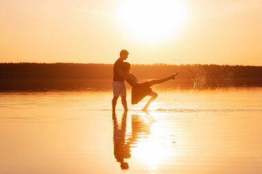 Silhouette of couple in love, dances with splashes in the water of the lake in the rays of orange sunset. Lovers hug each other, kiss. The man circles the woman in arms. Honeymoon travel of newlyweds