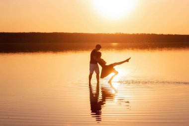 Silhouette of couple in love, dances with splashes in the water of the lake in the rays of orange sunset. Lovers hug each other, kiss. The man circles the woman in arms. Honeymoon travel of newlyweds