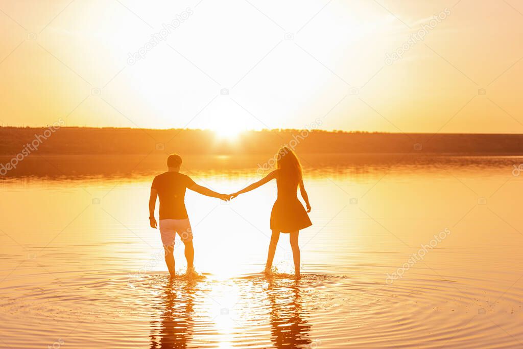 Silhouette of a couple in love. A man holds a woman's hand, walk on the water of the lake in the rays of the orange sunset. Lovers hug each other. Honeymoon travel of newlyweds. New life concept