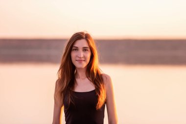 Gün batımında gökyüzüne doğru siyah bir elbise giymiş genç bir kadının yakın plan portresi. Kız saçı güneş ışınlarıyla aydınlatılır. Bir kadın gün batımında deniz kıyısında yürür. Yaşam tarzı seyahat hafta sonu