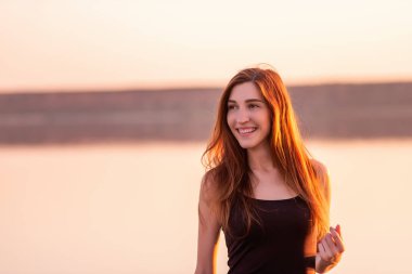 Close-up portrait of young woman in an airy black dress against the sunset sky. The girls hair is illuminated by the rays of the sun. A woman walks on the seashore at sunset. Lifestyle travel weekend