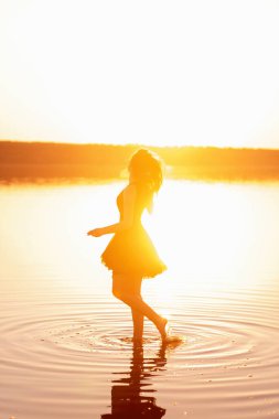 Gökyüzü yansıması olan gölün suyunda, havadar, siyah elbisesi olan genç bir kadının parlak, güneşli silueti. Kız gün batımında kıyıda denizde dans ediyor. Yaşam tarzı seyahati. Sağlık, hareket kolaylığı
