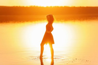 Gökyüzü yansıması olan gölün suyunda, havadar, siyah elbisesi olan genç bir kadının parlak, güneşli silueti. Kız gün batımında kıyıda denizde dans ediyor. Yaşam tarzı seyahati. Sağlık, hareket kolaylığı