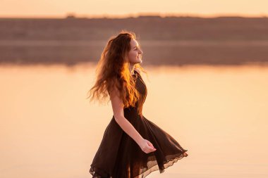 Kıvırcık, gün batımında havadar siyah elbiseli mutlu dansçının yakın plan portresi. Gölün sularındaki genç kadın deniz boyunca yürüyen gökyüzü yansımasıyla. Hayat tarzı seyahat hafta sonu. Sağlık, hareket kolaylığı