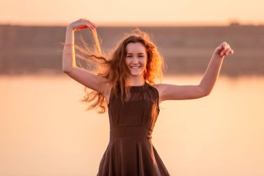 Kıvırcık, gün batımında havadar siyah elbiseli mutlu dansçının yakın plan portresi. Gölün sularındaki genç kadın deniz boyunca yürüyen gökyüzü yansımasıyla. Hayat tarzı seyahat hafta sonu. Sağlık, hareket kolaylığı