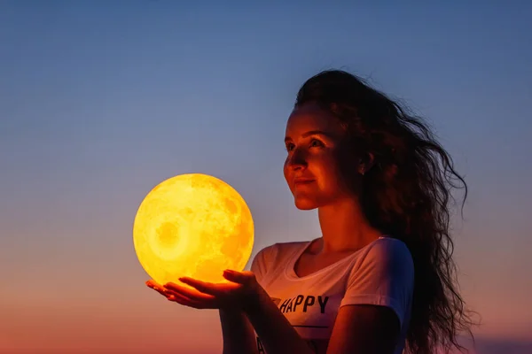 Genç bir kadın, kırmızı bir günbatımının arka planına karşı dolunayı elinde tutuyor. Milenyum blogcusu sosyal medyayı yönetiyor. Kız silueti portresi. Astroloji, Balmumu Hilal. Boşluğu kopyala