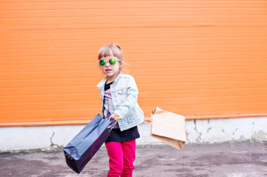 Little stylish girl in sunglasses with craft bags on shopping trip on an orange background. Shopaholic dances against the wall of the commercial center. Girl happy shopping on Black Friday. Copy space