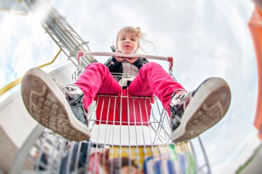 Küçük kız alışveriş arabasında otururken eğleniyor. Çocuk ailesiyle birlikte ticaret merkezine alışverişe gitti. Kız oynaşıyor, farklı duygular yaratıyor, dilini gösteriyor, at arabasına biniyor.