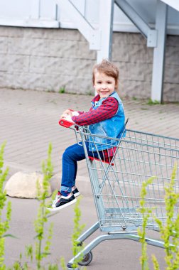 Küçük kız alışveriş arabasında otururken eğleniyor. Çocuk ailesiyle birlikte ticaret merkezine alışverişe gitti. Kız oynaşıyor, farklı duygular yaratıyor, dilini gösteriyor, at arabasına biniyor.