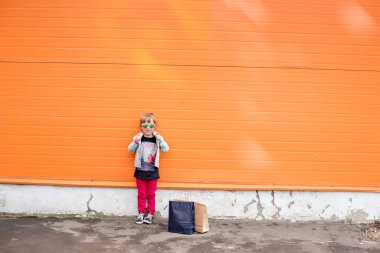 Little stylish girl in sunglasses with craft bags on shopping trip on an orange background. Shopaholic dances against the wall of the commercial center. Girl happy shopping on Black Friday. Copy space